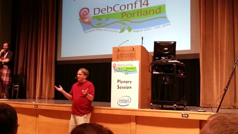 Linus Torvalds in a red shirt gestures on stage in front of a DebConf14 Portland slide and a lectern marked Plenary Session, with audience heads in the foreground.