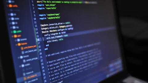 Close-up of a laptop screen in a dark room showing a code editor with a file tree on the left and numbered lines of Ruby test-configuration code in blue, green and orange.