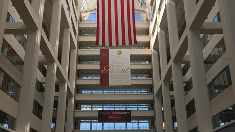 The tall white atrium of the US Patent and Trademark Office headquarters, lined with balconies, with a large American flag hanging from the skylight above a red-and-cream banner reading “Patent” and a smaller sign reading “Ten Million Patents”.