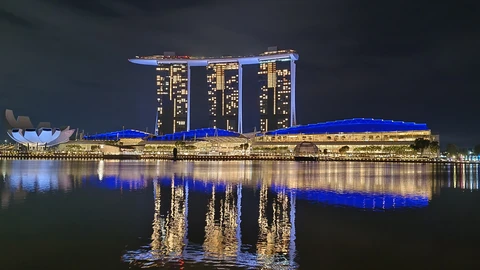 Night view across Marina Bay of the three Marina Bay Sands hotel towers joined by their rooftop SkyPark, the lit convention-centre roofline below them and the lotus-shaped ArtScience Museum at left, all reflected in the dark water.