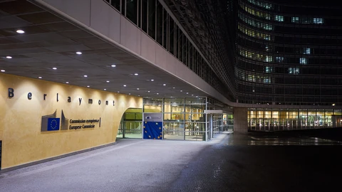 The entrance of the Berlaymont building in Brussels at night: a warmly lit wall with the word “Berlaymont” and the European Commission logo, a glass lobby, and rows of lit office windows rising above an empty forecourt.