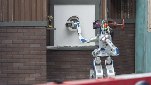 A white-and-blue bipedal humanoid robot with red accents stands at a wooden wall panel and uses a hand tool to cut a hole in drywall, next to a mounted valve wheel, on an outdoor competition course.