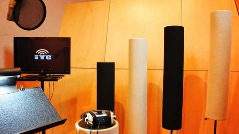 A vocal booth at In Your Ear Studios with cylindrical sound baffles, a music stand, a microphone stand and a flat-screen monitor, photographed from a low angle against wood-panel walls.