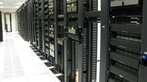 A long aisle of black network and server racks on a raised white floor under fluorescent ceiling lights, photographed looking down the row.