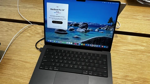 A Space Black 14-inch MacBook Pro with an M5 chip open on a light wooden store table, its screen showing a lake wallpaper and an Apple product card, cables running to the side.