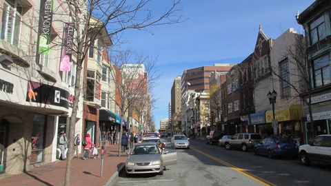 Market Street in downtown Wilmington, Delaware, on a sunny day: brick sidewalks, shopfronts and bare trees on both sides, parked cars, and office towers in the distance under a blue sky.