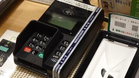 A black card payment terminal with a numeric keypad, small screen and contactless payment symbol on a shop counter, next to a white QR-code scanner and Japanese payment signs.