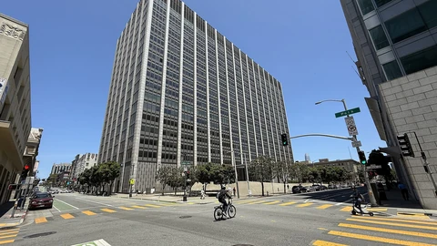 The tall, grid-windowed Phillip Burton Federal Building in San Francisco under a clear blue sky, seen across a wide intersection with yellow crosswalk stripes and a green bike lane.