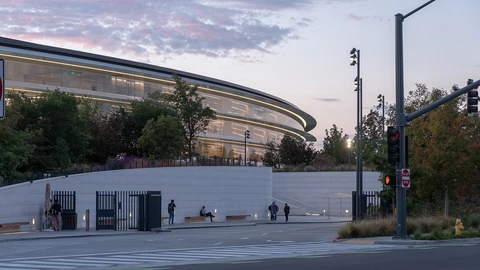 The long, curved glass facade of Apple Park’s ring building at dusk, seen behind a low white perimeter wall with a closed gate, trees and a streetlight under a pink and blue sky.
