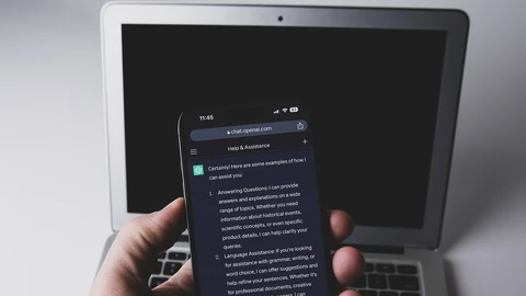 A hand holding a smartphone that shows a ChatGPT conversation, in front of an open silver laptop on a white desk.