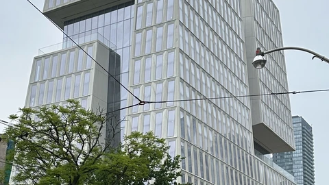 A tall, glass-clad building made of offset stacked blocks on a Toronto street corner, with trees, street lamps and an overcast sky.