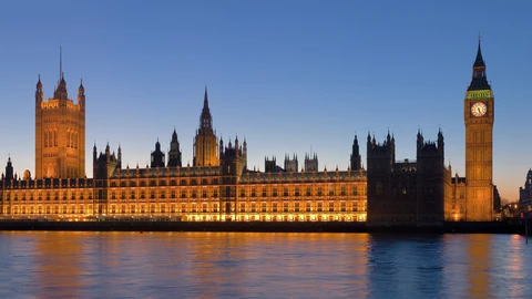 The Palace of Westminster lit up at dusk across the River Thames, with Victoria Tower on the left, Elizabeth Tower and its clock face on the right, and the floodlit facade reflected in the water.