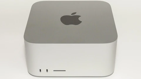 A compact silver Mac Studio desktop computer seen from the front on a reflective white surface, with a black power cable trailing behind it.