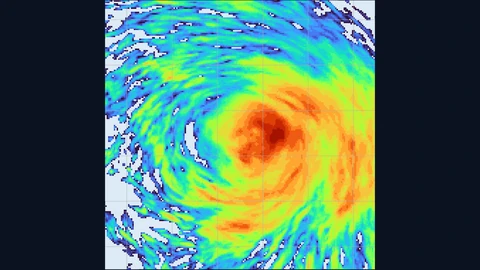 A simulated hurricane seen from above as a spiral of rainfall: a dark red core of heavy rain surrounded by orange, yellow and green rain bands curling outward, with pale dry patches at the edges, on a dark background.
