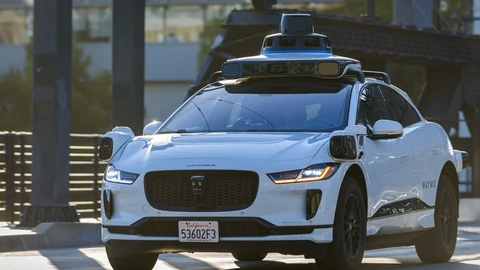 A white Waymo Jaguar I-PACE robotaxi with a roof-mounted lidar and camera dome drives toward the camera in low evening sun.