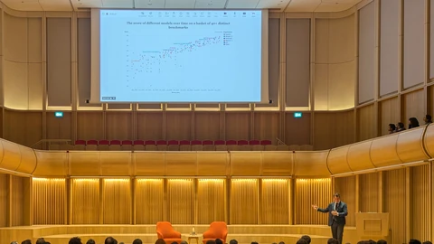 Jack Clark in a dark suit gestures on a wood-panelled concert-hall stage beside two orange armchairs, under a large projected chart titled The score of different models over time on a basket of 40+ distinct benchmarks, with an audience in the foreground.