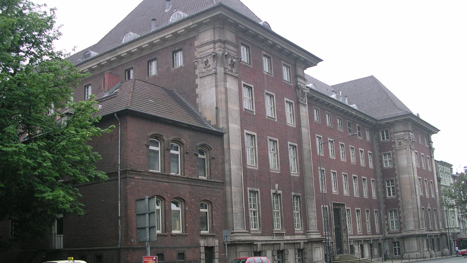 The red-brick and sandstone north front of the Aachen Regional Court building on Adalbertsteinweg, with tall windows, a steep slate roof and a leafy tree at the left edge.
