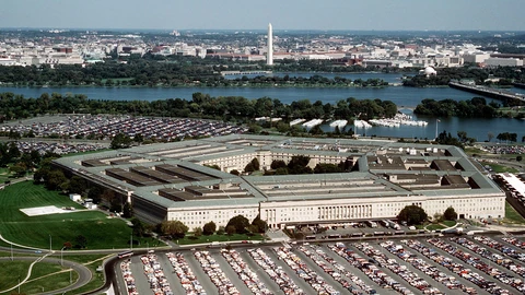 Aerial photo of the five-sided Pentagon building in Arlington, Virginia, with parking lots in front and the Potomac River and Washington Monument behind it.