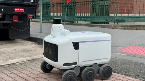 A white Avride sidewalk delivery robot with six black wheels, a dark front panel and a small sensor mast, standing on a red brick sidewalk beside a street.