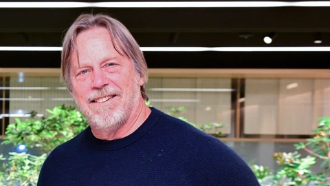 Tenstorrent CEO Jim Keller, smiling, with grey shoulder-length hair and a white beard, wearing a navy crew-neck sweater, standing in a bright lobby with green plants behind him.