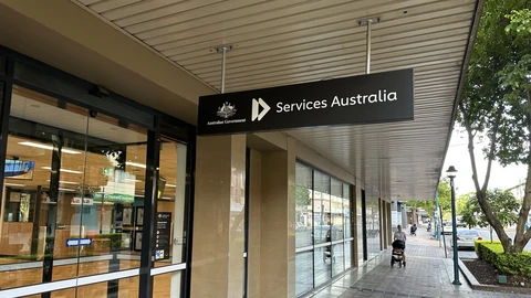 A black Services Australia sign with the Australian Government crest hanging under the awning of a glass-fronted service centre on a shopping street.