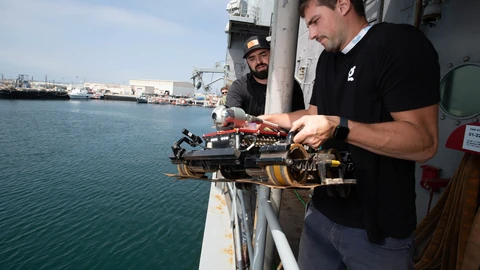 A Gecko Robotics engineer in a black T-shirt lowers a tracked inspection robot with magnetic wheels over the railing of a grey Navy test ship, with a colleague behind him and harbour water and piers in the background.