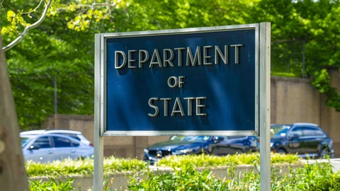 A dark blue sign with raised silver letters reading Department of State, on a metal frame in front of shrubs, a low wall, parked cars and green trees outside the Harry S. Truman Building in Washington.