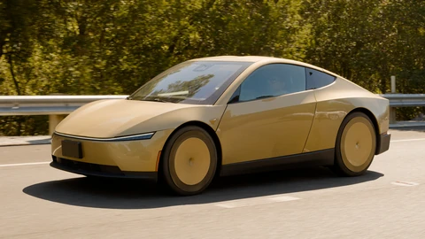 A gold two-door Tesla Cybercab with matching gold wheel covers driving on a California interstate in sunlight, seen from the front left, with a guardrail and trees behind it.