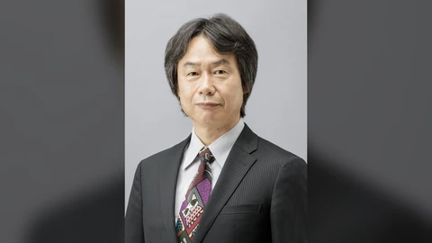 Official portrait of Shigeru Miyamoto in a dark pinstripe suit and patterned tie against a grey studio background, centred on a blurred, darkened copy of the same photo.