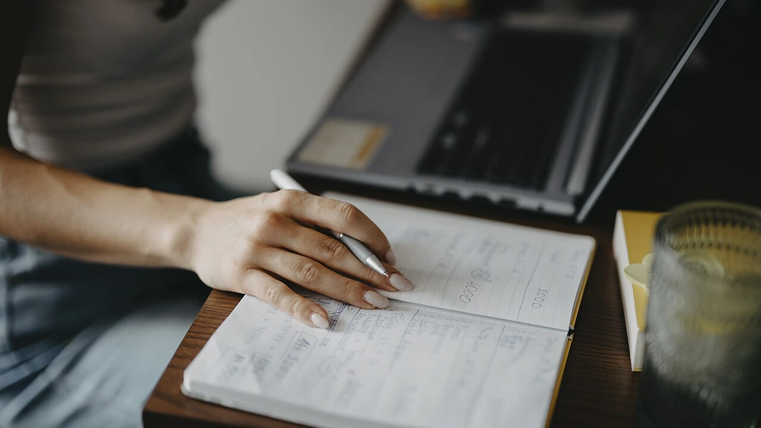 A person writing notes beside a laptop at a wooden desk.