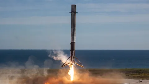 A SpaceX Falcon 9 first stage landing at Landing Zone 1 during the CRS-13 mission, trailing engines lit against a blue sky.